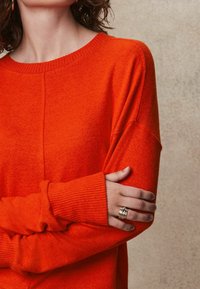 Oranger Strickpullover mit gerippten Bündchen, leicht oversized. Hautfarbe an den Händen sichtbar, geschmückt mit einem metallischen Ring. Neutraler Hintergrund.