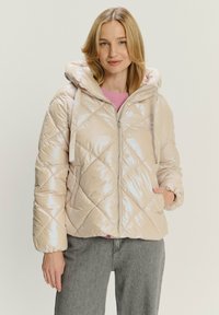Giacca imbottita e lucida di colore beige con un motivo a rombi, chiusura a cerniera e cappuccio. Presenta tasche laterali e polsini elasticizzati.