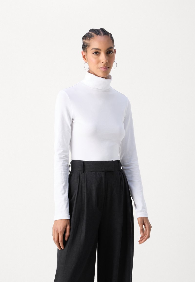 Maglietta bianca a maniche lunghe con collo alto dal design aderente, abbinata a pantaloni neri a vita alta e plissettati con una texture liscia e passanti per la cintura.