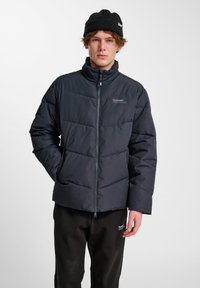 Mörk puffjacka med geometriskt quiltat mönster, dragkedja fram, hög krage och logodetalj. Bärs med svarta byxor och stickad mössa.