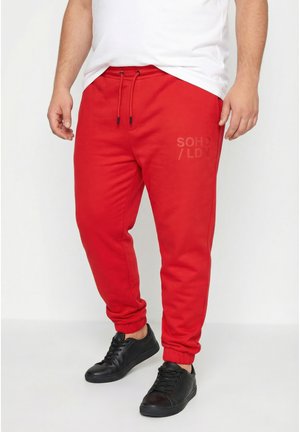 Mann trägt rote Jogginghosen mit Kordelzugbund und schwarzen Turnschuhen, kombiniert mit einem weißen T-Shirt, stehend vor einem einfarbigen Hintergrund.