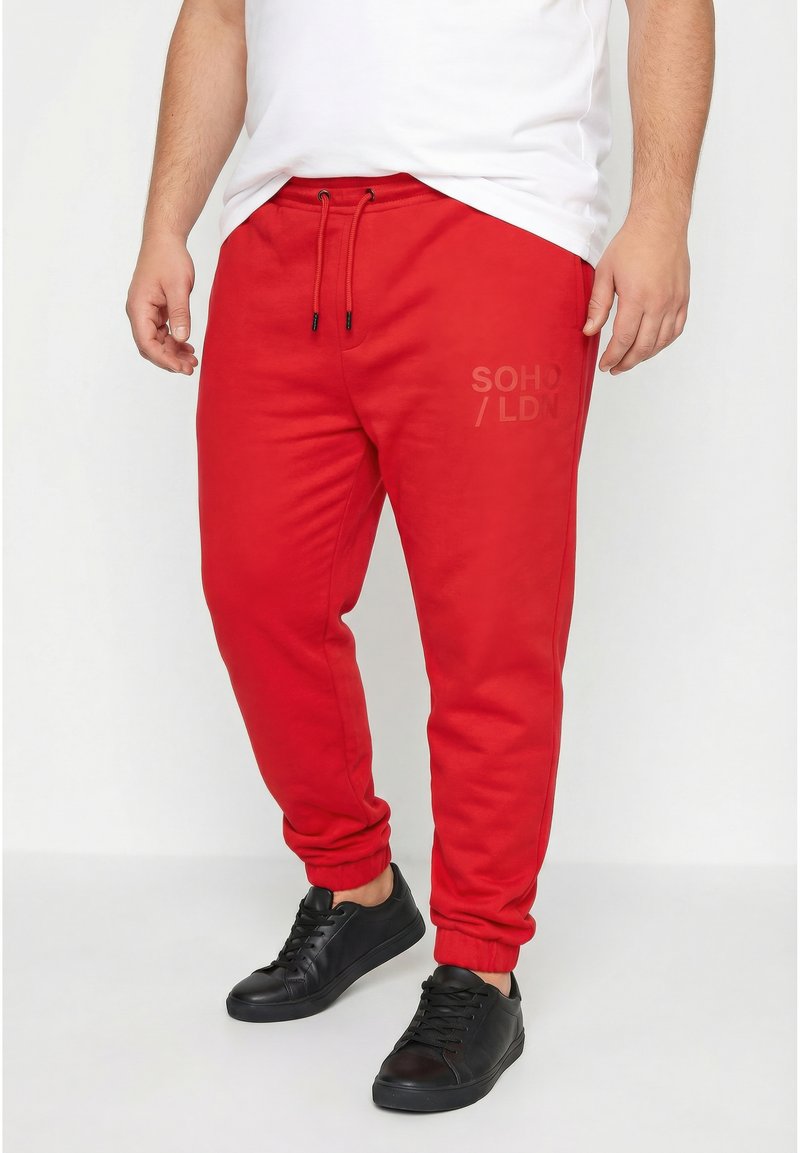 Mężczyzna w czerwonych spodniach dresowych ze ściągaczem w pasie i czarnych trampkach, ubrany w białą koszulkę, stoi na jednolitym tle.