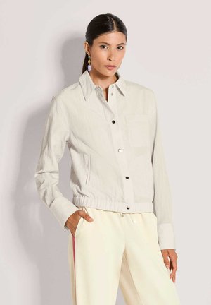 Hellgraue Hemdjacke mit Brusttasche, elastischem Bund und glatter Textur, kombiniert mit cremefarbenen Hosen mit rosa Seitenstreifen.