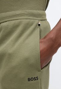 Κοντινό πλάνο ενός χεριού στην πλαϊνή τσέπη παντελονιού σε ελιά πράσινη υφή, με το λογότυπο "BOSS" κεντημένο κοντά στην τσέπη.