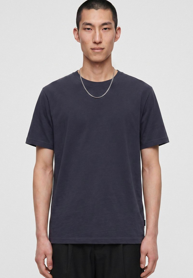 Homme aux cheveux courts portant un t-shirt gris foncé uni et un collier chaîne argenté, debout devant un fond gris clair.
