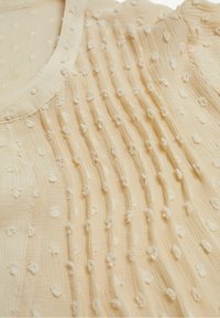 Beige fabric with textured raised dots and vertical pleats near a round neckline.