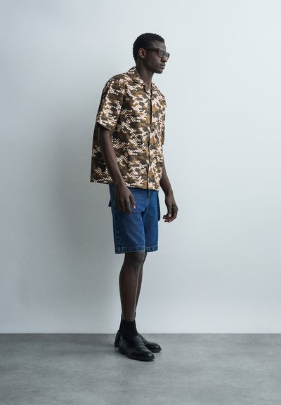 Άνδρας που φορά καφέ πουκάμισο με σύντομο μανίκι και σχέδια, μπλε τζιν σορτς μέχρι το γόνατο, μαύρες κάλτσες, μαύρα δερμάτινα παπούτσια και σκουρόχρωμα γυαλιά ηλίου, στέκεται σε εσωτερικό χώρο.