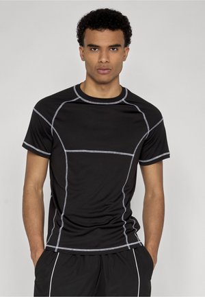 Mand iført en tætsiddende sort atletisk trøje med hvide syninger og sorte shorts med hvid piping, stående med hænderne i lommerne.