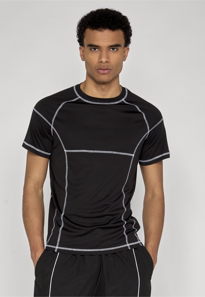 Homme portant un t-shirt de sport noir ajusté avec des coutures blanches et un short noir avec un liseré blanc, debout les mains dans les poches.