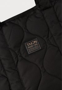 Μαύρη επενδεδυμένη τσάντα με υφή, που διαθέτει ένα ορθογώνιο ετικέτα με χρυσά γράμματα που περιλαμβάνει το όνομα της μάρκας "TAION."