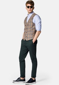 Brązowa kamizelka w kratę nałożona na jasnoniebieską koszulę, w połączeniu z ciemnozielonymi spodniami i granatowymi sneakersami. Kamizelka ma podwójnie zapięty krój oraz guziki.