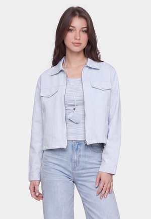 Junge Frau mit langen braunen Haaren trägt eine hellblaue Jacke, ein gestreiftes Shirt, hellblaue Jeans und einen durchsichtigen Ausweis an einem Band um den Hals.