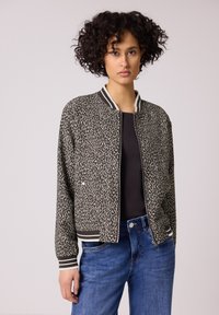 Donna con capelli ricci che indossa una giacca bomber nera e beige con stampa leopardata, una maglietta nera e jeans blu su uno sfondo neutro.