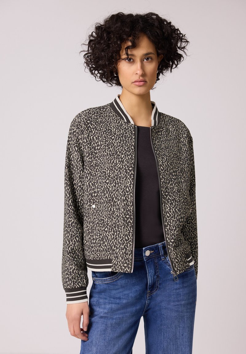 Donna con capelli ricci che indossa una giacca bomber nera e beige con stampa leopardata, una maglietta nera e jeans blu su uno sfondo neutro.