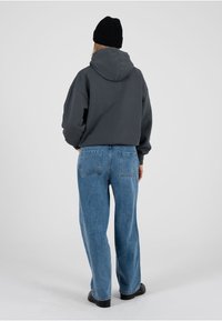 Grauer Kapuzenpullover mit großer Kapuze, kombiniert mit hellblauen, weit geschnittenen Jeans. Das Model trägt eine schwarze Strickmütze und schwarze Schuhe.