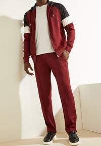 Chaqueta deportiva roja con cremallera, con detalles en negro y blanco, combinada con pantalones a juego. Tejido suave, bolsillos laterales y zapatillas negras.