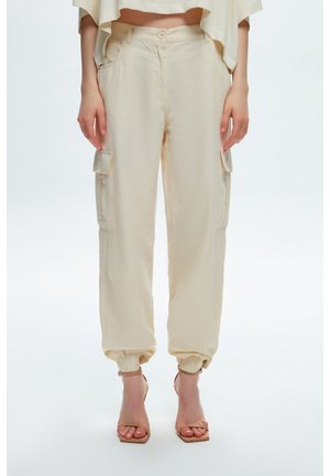 Femme portant un pantalon style cargo beige avec poignets élastiques et des sandales à talons ouverts beiges, debout sur fond blanc.