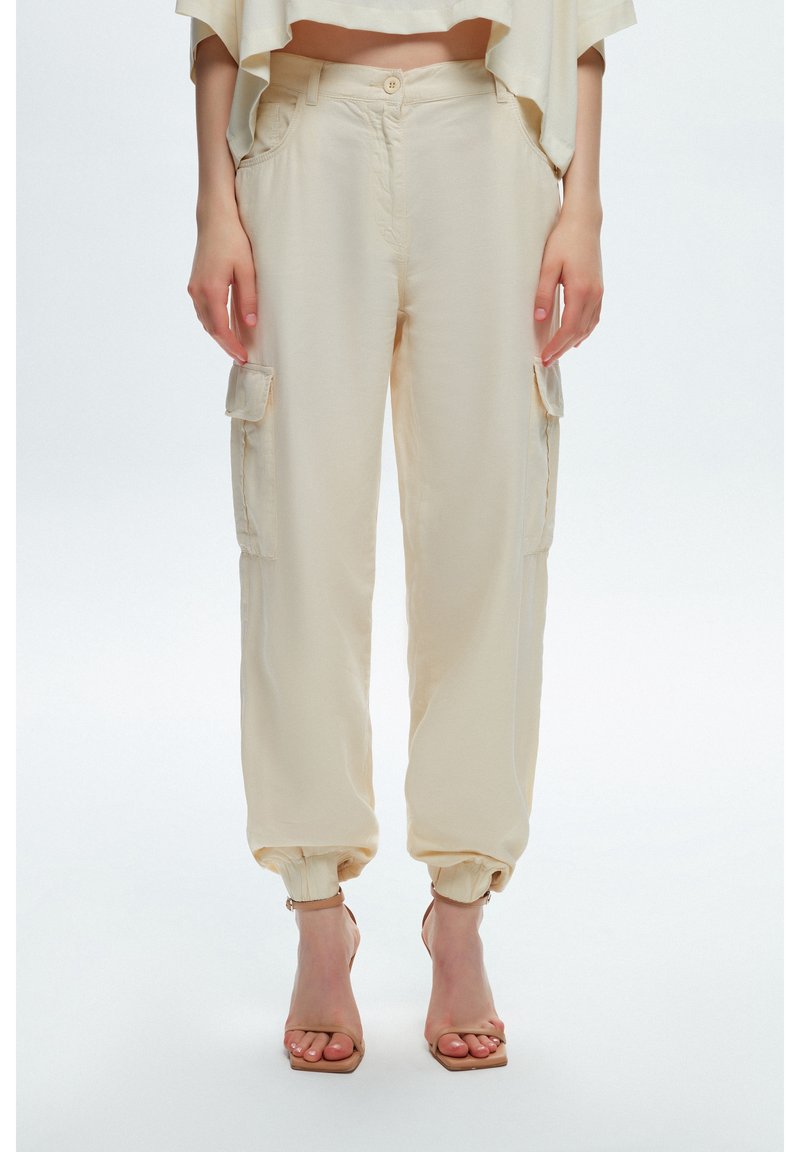 Femme portant un pantalon style cargo beige avec poignets élastiques et des sandales à talons ouverts beiges, debout sur fond blanc.