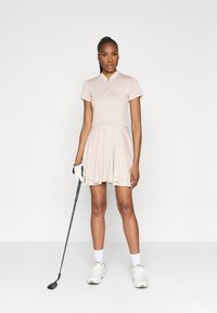 Polo-Kleid in hellbeige mit einem Reißverschluss-Kragen, kurzen Ärmeln und einem ausladenden Rock. Accessories sind ein Golfclub und ein Handschuh.