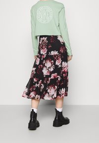 Mátově zelený zkrácený top s dlouhým rukávem a bílým logem, v kombinaci s černou květinovou maxi sukni se volány a růžovými, bílými a burgundskými květinami, nošený s černými kotníkovými botami.