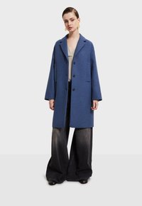 Modrá vlnená bunda s veľkými náprsnými vreckami, s vykrojeným golierom a čiernymi gombíkmi, nosená cez sivé voľné deninové nohavice.