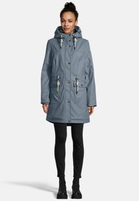 Woman wearing a dusty blue hooded raincoat with button and rope details, black leggings, and black lace-up boots, standing against white background.