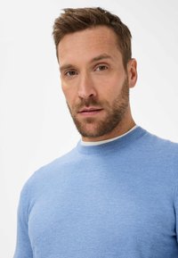 Helles blaues Strickpullover mit Rundhalsausschnitt, feiner Textur und dezenten Rippen am Kragen. Über einer weißen Schicht getragen.