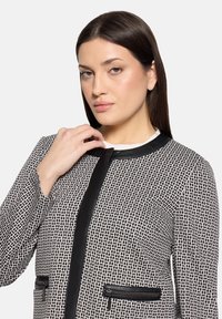 Veste à motifs noir et blanc en tissu texturé, avec un col noir contrastant et des poches zippées.