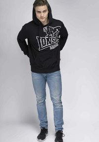 Sudadera negra con un gráfico de león blanco y el texto "LONDRES", combinada con jeans de color azul claro y zapatillas negras. El modelo está de pie con las manos en los bolsillos.