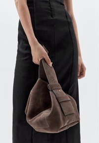 Brown suede handbag with a thick strap, featuring a structured shape and contrasting stitching. Held by a person wearing a black dress.