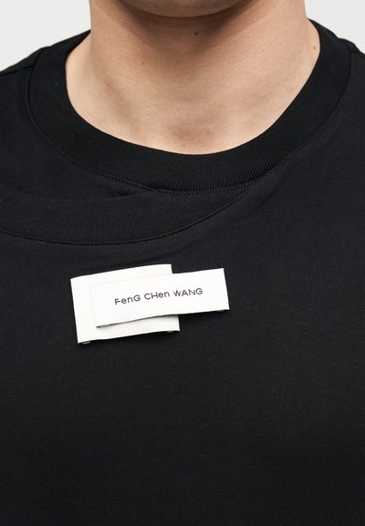 Κοντινό πλάνο ενός ατόμου που φοράει μαύρο μπλουζάκι με στρογγυλή λαιμόκοψη και μια υφασμάτινη ετικέτα ραμμένη στο στήθος με την επιγραφή "FenG CHen WANG."