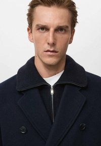 Cappotto di lana blu navy con colletto a coste e chiusura con zip, caratterizzato da bottoni doppi e una superficie liscia. Strato interno bianco visibile.