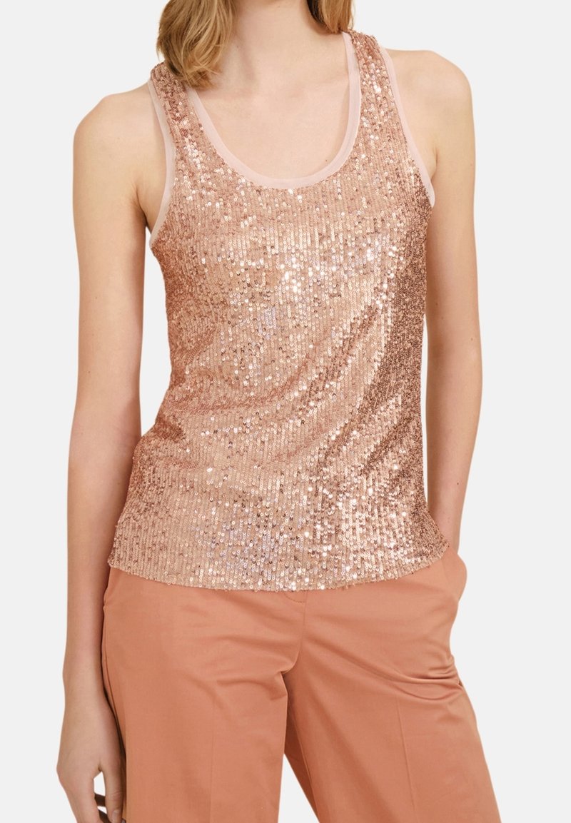 Femme portant un débardeur sans manches en sequins rose doré et un pantalon taille haute couleur pêche, debout avec une main dans la poche sur fond blanc.