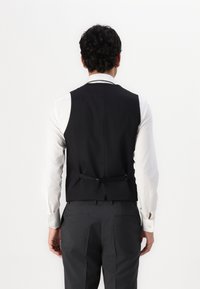 Schwarze Weste mit taillierter Passform, ausgestattet mit einem geschichteten Rückendesign und verstellbaren Trägern, getragen über einem weißen Hemd und kombiniert mit grauen Hosen.