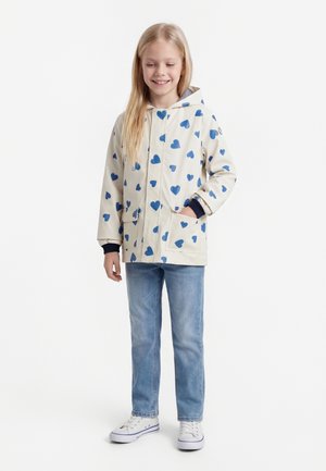 Jauna mergaitė šypsosi, dėvi kreminę gobtuvą su mėlynomis širdelėmis, šviesiai mėlynas džinsus ir baltus sportbačius, stovinti priešais baltą foną.