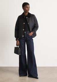 Black quilted jacket with gold buttons, paired with dark blue flared jeans. Holding a small black handbag, standing on a neutral surface.