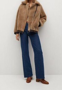 Brązowa kurtka z sztucznego futra z zamkiem z przodu, szerokim kołnierzem i kontrastującymi czarnymi detalami, zestawiona z niebieskimi dżinsami o wysokim stanem i brązowymi botkami za kostkę.