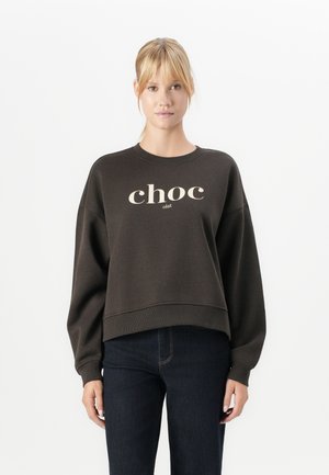 Vrouw met blond haar in een oversized donkerbruine sweatshirt met de tekst "choc olat" en een donkerblauwe spijkerbroek tegen een witte achtergrond.
