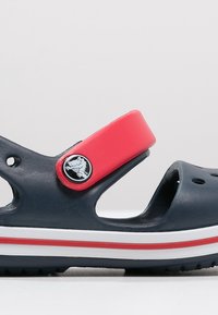 Navy blue sandal made of rubber with a red adjustable strap, white accents, and ventilation holes. Features a circular logo on the strap.
