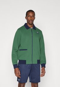 Casaco verde com relevo, gola azul-marinho, fecho de correr e dois bolsos frontais. Logotipo bordado no peito. Combinado com calções azul-marinho.