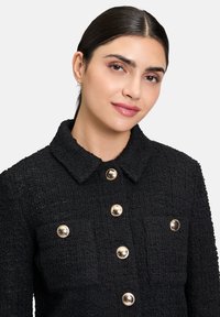 Giacca nera con texture, colletto, bottoni dorati e due tasche frontali. Presenta un tessuto intrecciato per una maggiore profondità e dimensione.