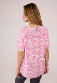 Kurzärmlige Bluse mit pink-weißer Blumenmuster, die einen abgerundeten Rückensaum und einen knöpfbaren Schlüssellochverschluss am Halsbereich aufweist.
