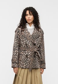 Καμπαρντίνα με λεοπάρ σχέδιο σε μπεζ και μαύρο, διπλό στήθος με ζώνη στη μέση, μεγάλοι γιακάδες και μανσέτες με κουμπιά, φορεμένη πάνω από γκρι πουλόβερ.