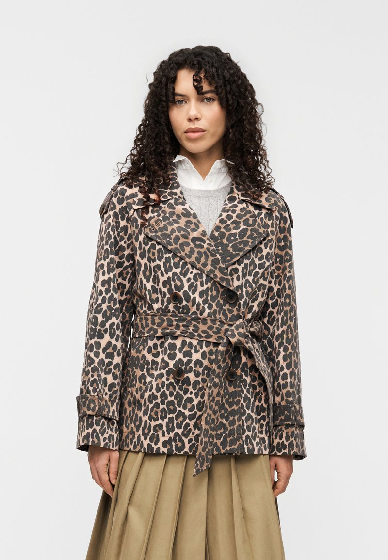 Καμπαρντίνα με λεοπάρ σχέδιο σε μπεζ και μαύρο, διπλό στήθος με ζώνη στη μέση, μεγάλοι γιακάδες και μανσέτες με κουμπιά, φορεμένη πάνω από γκρι πουλόβερ.