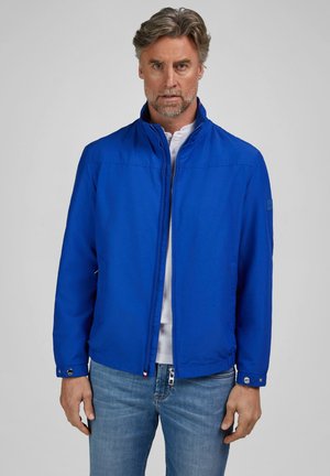 Man met grijs haar en baard die een felblauwe jas met rits draagt over een wit shirt en blauwe spijkerbroek, staand tegen een effen achtergrond.