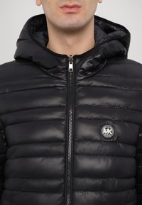 Chaqueta de plumón negra con capucha, que presenta un diseño acolchado, cierre frontal con cremallera y un logotipo circular plateado en el lado izquierdo del pecho.