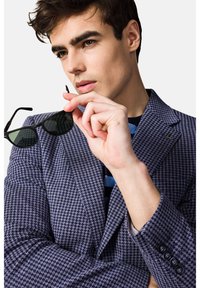 Granatowa marynarka w wzór houndstooth o dopasowanym kroju. Model trzyma czarne okulary przeciwsłoneczne. Pod marynarką widoczna niebieska i czarna koszula w paski.