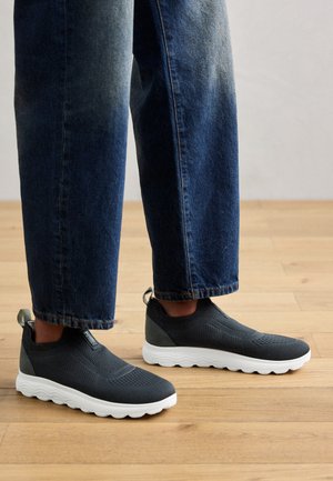 Zapatillas negras de malla sin cordones con suelas de goma blanca, que presentan un upper texturizado, una silueta elegante y una lengüeta en el talón.