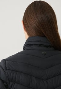 Sort dunjakke med høj krave, udstyret med et chevron quiltet mønster og et logo på ryggen nær halsen. Glat tekstur.