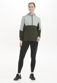 Letvægts zip-up jakke i lys grå og olivengrøn, med hætte, elastiske manchetter og et strømlinet design. Båret sammen med sorte leggings.